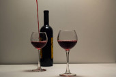 Dvě sklenice naplněné červeným vínem a do jedné sklenice se nalije nápoj. Láhev červeného vína, jednu naplněnou sklenici, druhou sklenici naplněnou, na lehkém podkladu, s lehkým pozadím.