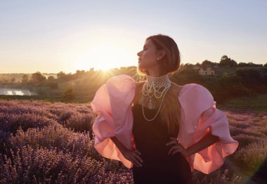 Güzel bir kadın lavanta tarlasında gün batımında siyah ve pembe elbiseler içinde dinleniyor. Çiçek tarlasındaki sarışın kadın. Provence.