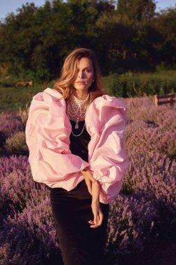 Güzel bir kadın lavanta tarlasında gün batımında siyah ve pembe elbiseler içinde dinleniyor. Çiçek tarlasındaki sarışın kadın. Provence.