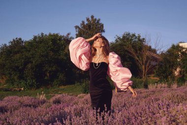Güzel bir kadın lavanta tarlasında gün batımında siyah ve pembe elbiseler içinde dinleniyor. Çiçek tarlasındaki sarışın kadın. Provence.