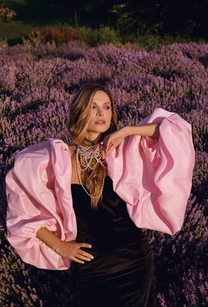 Güzel bir kadın lavanta tarlasında gün batımında siyah ve pembe elbiseler içinde dinleniyor. Çiçek tarlasındaki sarışın kadın. Provence.
