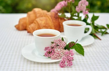 İki fincan siyah çay, çiçekler ve beyaz arka planda taze kruvasanlar. Bahar kahvaltısı kavramsal kompozisyonu