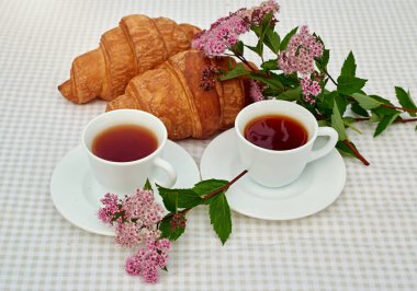 İki fincan siyah çay, çiçekler ve beyaz arka planda taze kruvasanlar. Bahar kahvaltısı kavramsal kompozisyonu