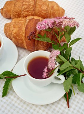 İki fincan siyah çay, çiçekler ve beyaz arka planda taze kruvasanlar. Bahar kahvaltısı kavramsal kompozisyonu