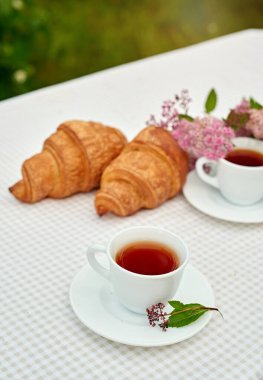 İki fincan siyah çay, çiçekler ve beyaz arka planda taze kruvasanlar. Bahar kahvaltısı kavramsal kompozisyonu