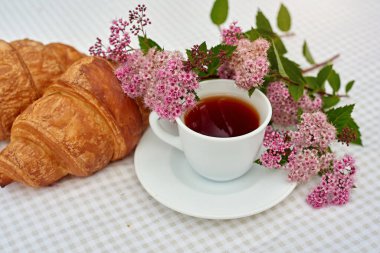 İki fincan siyah çay, çiçekler ve beyaz arka planda taze kruvasanlar. Bahar kahvaltısı kavramsal kompozisyonu