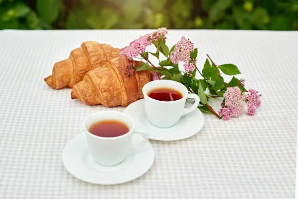 İki fincan siyah çay, çiçekler ve beyaz arka planda taze kruvasanlar. Bahar kahvaltısı kavramsal kompozisyonu