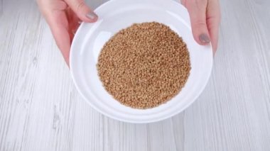 Kadının elleri masaya karabuğday koyuyor. Sağlıklı yiyecekler. Beyaz tabakta karabuğday taneleri. Sağlıklı tahıllar