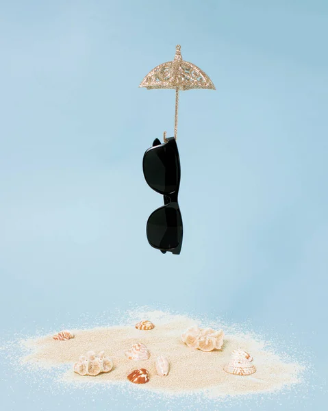 Falling sunglass hanged on umbrella on bright sea sand decorated with shells on pastel blue background. Minimal summer vibe, party theme concept. Idyllic tropical holiday.