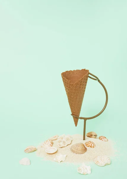 Ice cream cone on stand underneath decorated with sand and seashells on pastel green background. Minimal summer, vacation concept. Create layout with copy space.