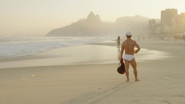 イパネマビーチに立っている男を通り過ぎるスローモーショントラッキングショット 男と女が波に向かって歩く ブラジル リオデジャネイロで撮影 — ストック動画