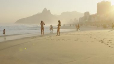 Rio De Janeiro, Brezilya - Haziran 2013: Yavaş çekim, Dois Irmaos ve alacakaranlıkta şehir manzarası arka plan karşı Ipanema plajda bikinili iki genç kadın izleme çekim