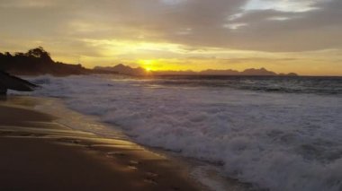 Yavaş çekim, ufukta dağlar ile Ipanema sahilde kayalara karşı gün batımında gelgit rulo olarak dolly dışarı. Arka planda gökyüzünde bulutlar ve şehir manzarası