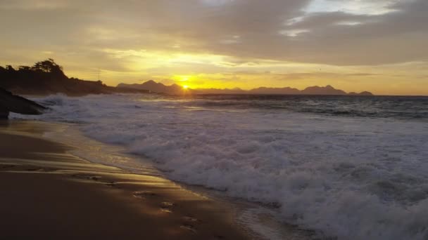 慢动作 当潮水在日落时分向伊帕内马海滩上的岩石上滚滚而来 山在地平线上时 娃娃滚出来 天空中的云彩和背景中的城市景观 — 图库视频影像