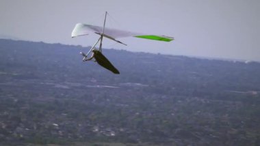 Bir askı planörü havada süzülürken görülür. Uzaktaki dağların ve vadinin manzarası. Bir yamaç paraşütü kısaca görülür.