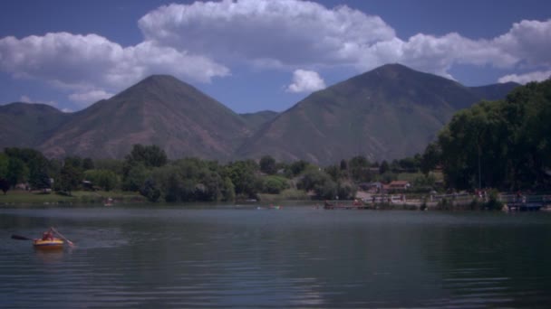 两个人在湖上划着木筏的镜头 它们位于框架的左下角 被群山和树木包围着 也有人沿着框架右侧的码头行走 — 图库视频影像