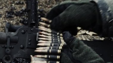 Eldivenli ellerin, kemerle beslenen bir makineli tüfeğin besleme tepsisine bir mermi zinciri yüklemesinin yakın çekimi. Daha sonra kapatılır ve o ateş etmeye hazırlanırken askerlerin yüzlerini görüruz. Yeşil Bereli Abd Ordusu Özel Kuvvetler için bir eğitim gönderen.