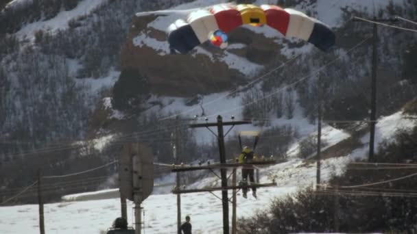 雪の山道でスキーに降りて着陸するベースジャンパーのスローモーションショット 彼と彼の友人は勝利に腕を上げる 送電線が近くにあり 背の高い山が背景にある — ストック動画