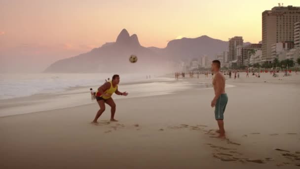 里约热内卢 2013年6月 慢动作锅 在里约热内卢伊帕内马海滩的两个男人在日落时分踢足球 或足球 对维迪加尔的天际线 — 图库视频影像