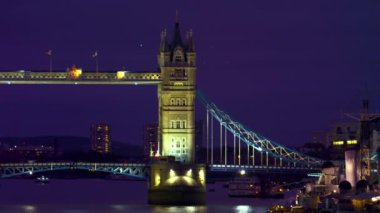 Londra'da akşam aydınlatılmış Tower Bridge bir kaydırma atış - sağdan sola - . Köprüde trafik, suda tekneler ve arka planda bazı binalar var..