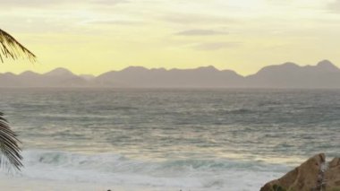 Rio de Janeiro, Brezilya'da Ipanema plaj dalgaları ve tepeler üzerinde gün batımı panning çekim. Bir palmiye ağacı çekim foregrounds.