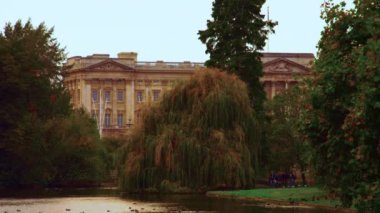 Buckingham Sarayı'nın, Saint James Park'tan çekilen arka planda ki sabit çekimi. Parkta yürüyen bir sürü ağaç, bir su yolu ve bazı insanlar var..
