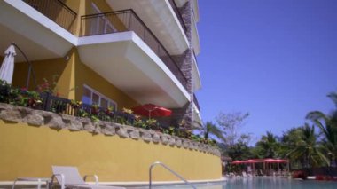 Balkonlu bir tatil köyü binasına bakan bir panning çekim. Atış açıları binanın dışındaki mavi havuza kadar iner..
