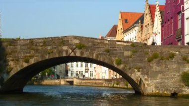 Bruges, Belçika'da bir kanalın üzerinde taş köprünün altından geçiyor. Kanalın yanında renkli binalar ve köprüden geçen bir adam var..