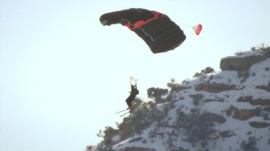 Bir baz atlayıcısı kayalık çıkıntıdan atlamış gibi görünüyor. O sola yönelir sonra dağlar arkasında görünümüne gelir gibi sağa döner. Bu yavaş çekimde çekilir..