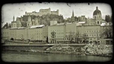 Salzburg'ın eski şehir merkezinde geniş atış, bitişik bir büyük nehir ve ön planda soğuk çim; eski tarihi binaların meşgul sokaklarda satır ve şehrin ünlü Festung Hohensalzburg Kalesi manzaralıdır