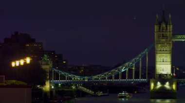 Londra'da akşam aydınlatılmış Tower Bridge bir kaydırma atış - soldan sağa - . Köprüde trafik, suda tekneler ve arka planda bazı binalar var..