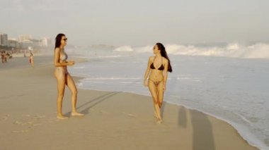 Rio De Janeiro, Brezilya - Haziran 2013: Yavaş çekim, güneş gözlüğü takan bikiniler içinde Ipanema plajında iki kızın fotoğrafını takip etmek, kumda gülmek ve gülümsemek. Kameranın gölgesi kumda görülebilir.