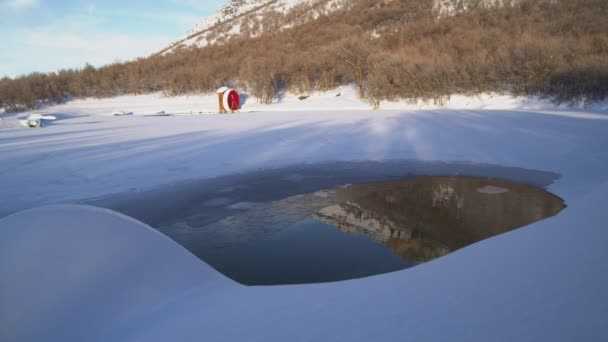 雪に覆われた凍った湖の端に赤い水車の時間経過 茶色の丘がプールの端まで流れ落ち 水の非Fozenパッチに反射します — ストック動画