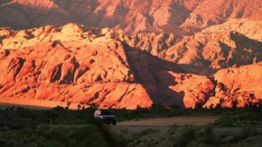 Öğleden sonra bir çöl karayolu üzerinde güneşli kırmızı tepelerin önünden geçen araba açılı görüntüleri. Çöl bitkileri yol çevresinde görülebilir. Nevada'da çekildi.