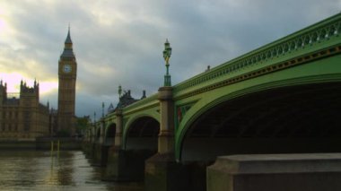 Sağdaki Westminster Köprüsü'nden başlayarak, kamera ön planda Thames Nehri ile Westminster Sarayı ve Big Ben panoramik görünümü üzerinde süzülür.