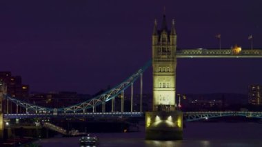Bir kaydırma atış - soldan sağa - aydınlatılmış Tower Bridge (Londra'da) akşam. Köprüde trafik, suda tekneler ve arka planda bazı binalar var..