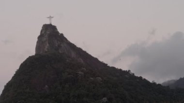 Rio de Janeiro'daki Corcovado'daki İsa heykelinin statik bir fotoğrafı. Heykel çerçevenin sol üst kısmında. Bir manzara kuşları aşağıdaki uzak tepelerde uçuyorlar. Gökyüzü bulutlarla dolu..