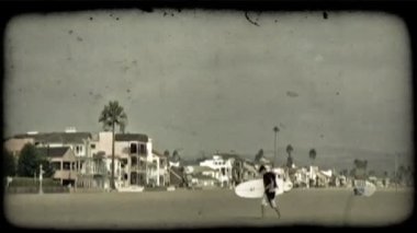 Genç adam surfer sörf tahtası, taşıyan Güney Kaliforniya kumsala evler, kınamak ve bulutlu gökyüzü altında arka planda beach kiralama ile birlikte yürür. Vintage stilize video klip.