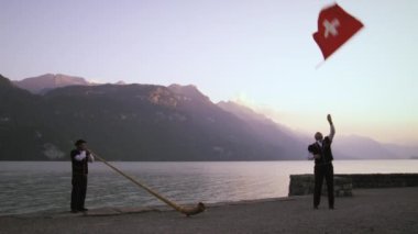 Adam alphorn oyuncu yakınında dururken İsviçre bayrağı twirls ve atar. Her ikisi de arka planda dağlar olan bir gölün yanında duruyor, ve bir sandalda biri tarafından sırayla. İsviçre'de çekildi.