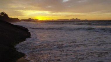 Yavaş çekim, praia do Diabo (Devil's Beach) gün batımında plaj ve kayalıklara isabet katil dalgaların statik çekim, arka planda altın ufuk
