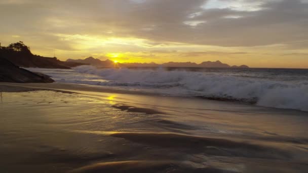 スローモーション 波が日没時にイパネマビーチに衝突するようにドリーアウト 地平線上の山々と空の雲 黄金色 — ストック動画