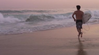 Gün batımında Ipanema sahilinde bir dalga gelirken sörf tahtasına atlayan, okyanusa koşan bir erkek sörfçünün yavaş çekim izleme çekimi