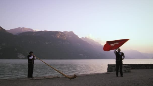 男子旋转和折腾瑞士国旗 而站在附近的阿尔卑斯球员 他们都站在一个湖边 背景是群山 在瑞士拍摄 — 图库视频影像
