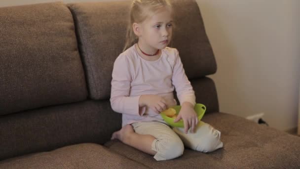 Liten flicka tittar på TV och äter chips sitter på soffan — Stockvideo