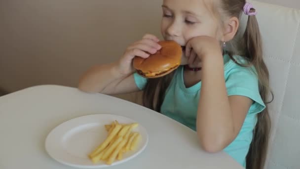 Kindheits- und Esskonzept - kleines Mädchen genießt Burger und Pommes auf dem Tisch und genießt ungesundes Essen — Stockvideo