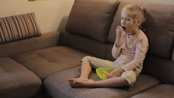 Девочка смотрит телевизор и ест чипсы, сидя на диване — стоковое видео