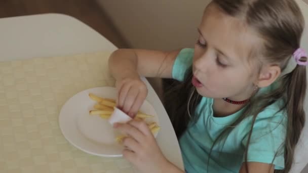 En liten flicka som sitter vid bordet och äter pommes frites med sås. Flicka klädd i blå t-shirt — Stockvideo