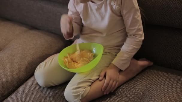 Dziewczynka ogląda telewizję i je chipsy siedząc na kanapie — Wideo stockowe