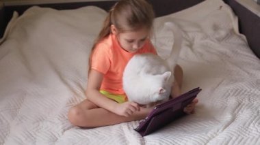 Küçük kız çevrimiçi oyunlarda tablette oynar beyaz kedinin yanında yatar.