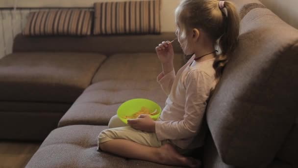 Fetiță uitându-se la televizor și mâncând chips-uri stând pe canapea — Videoclip de stoc